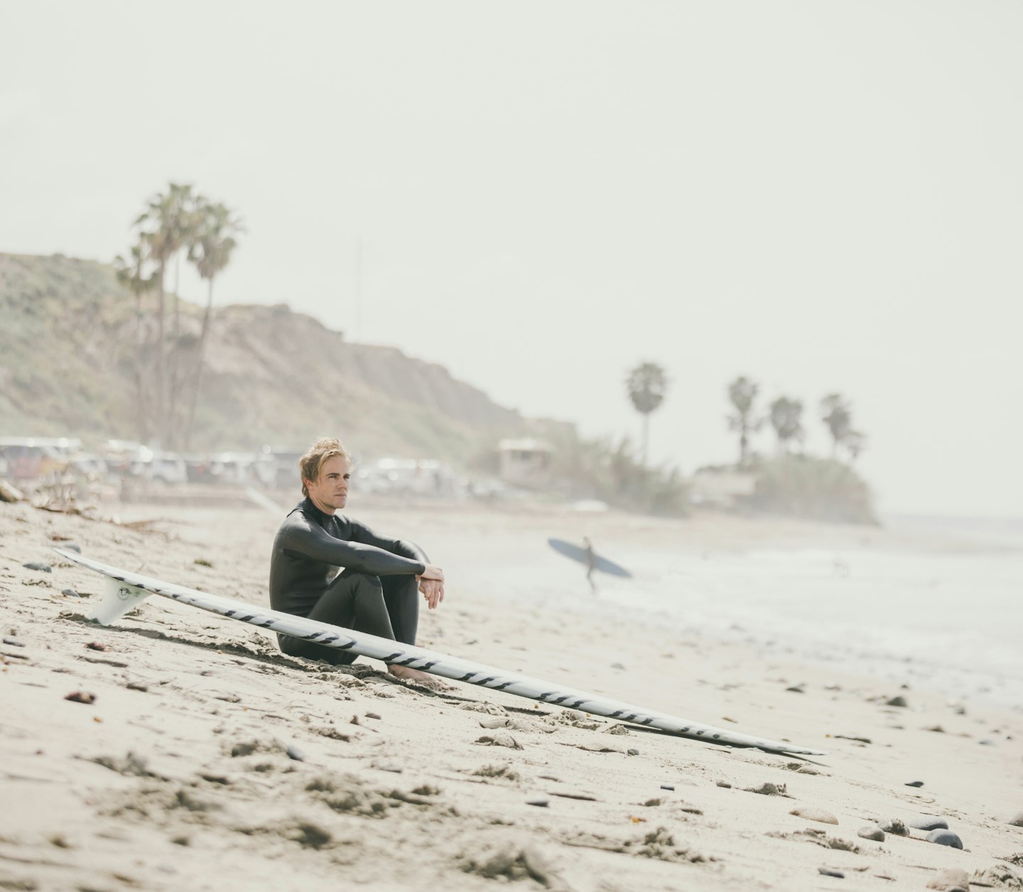 A man sitting on the beach next to his surfboard, looking into the distance.
